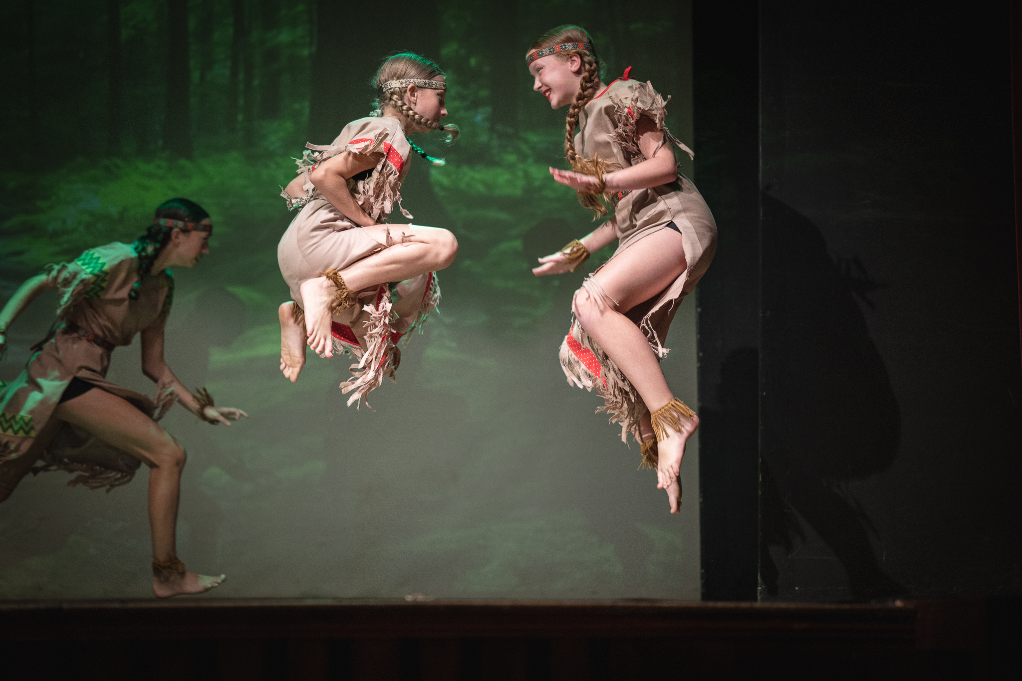 Two young female performers in Indigenous-inspired fringed costumes mid-air in a synchronized jump on a stage, facing each other with smiles. A third performer is visible in the background against a forest backdrop.